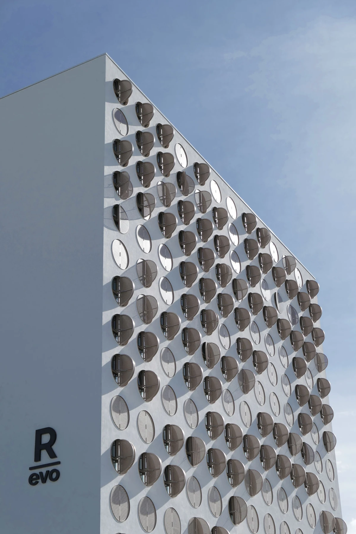 White modern building facade dotted with circular sunshades in a grid, set against a clear blue sky.