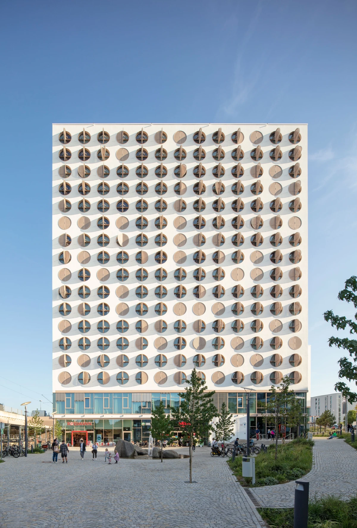 Modern high-rise building with circular balconies arranged in a grid, facing a cobblestone plaza with trees and pedestrians.
