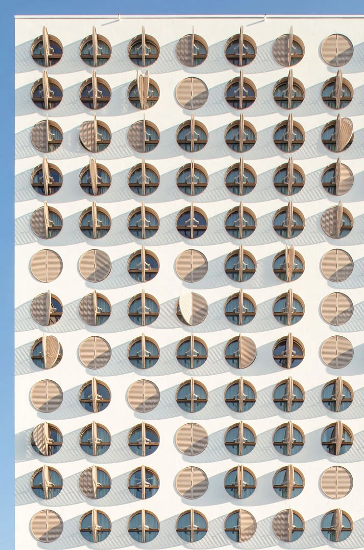 White building facade with a grid of circular brown-framed windows with vertical slats, casting diagonal shadows.