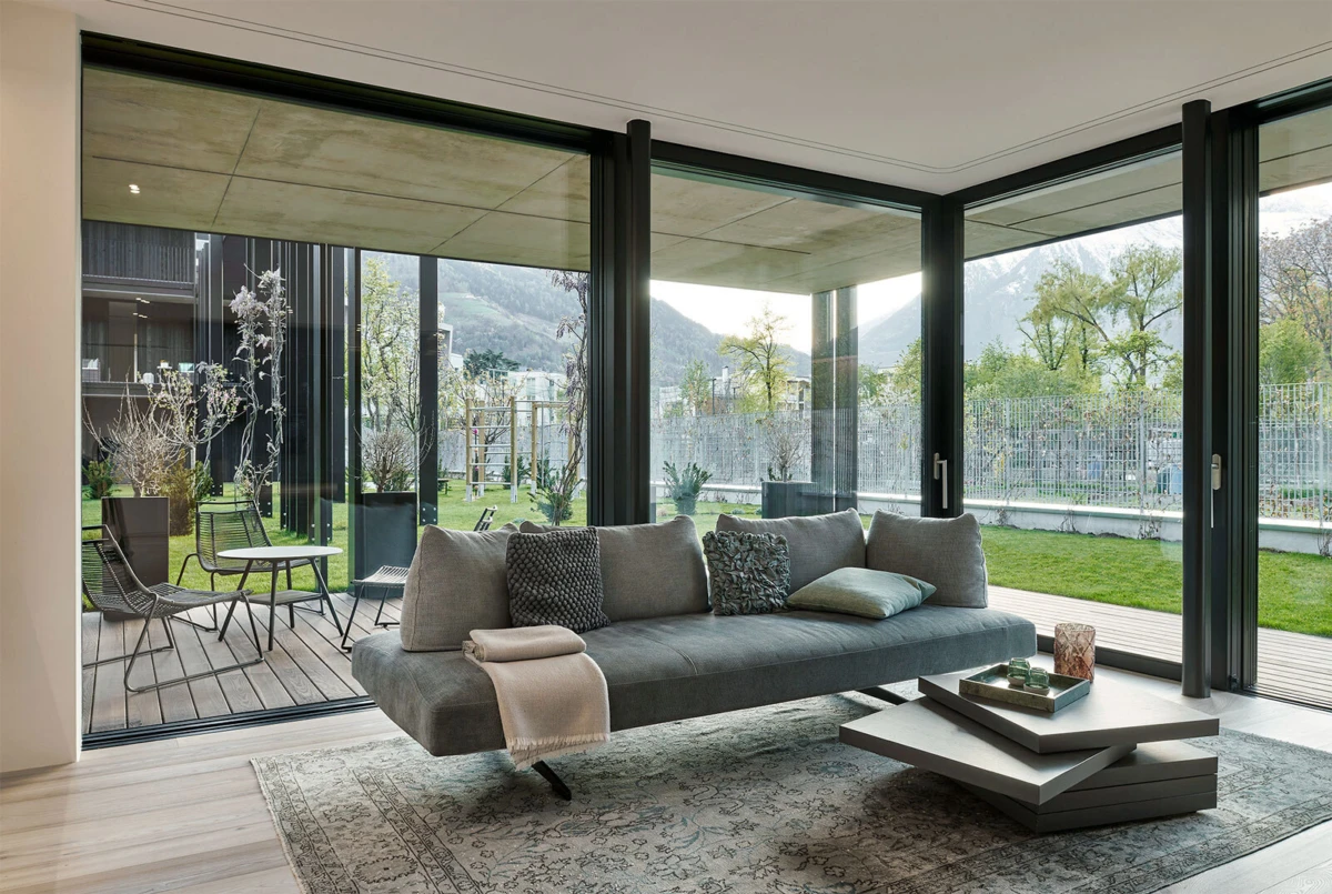 Gray sectional sofa with cushions in a bright living room with floor-to-ceiling glass walls opening to a wooden deck and garden.