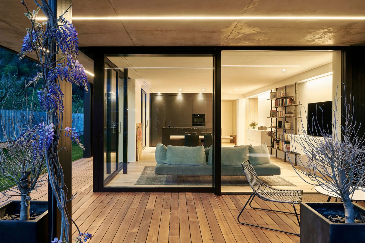 Open-plan living room visible through glass sliding doors; teal sofa, bookshelf, kitchen island; wooden deck with purple wisteria planter.