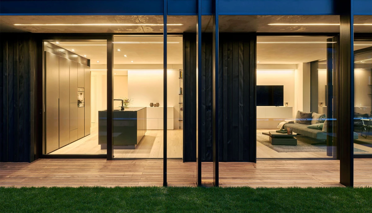 Open-plan kitchen and living room viewed through large glass walls at night; dark island, built-in appliances, sofa, and TV; wooden deck outside.