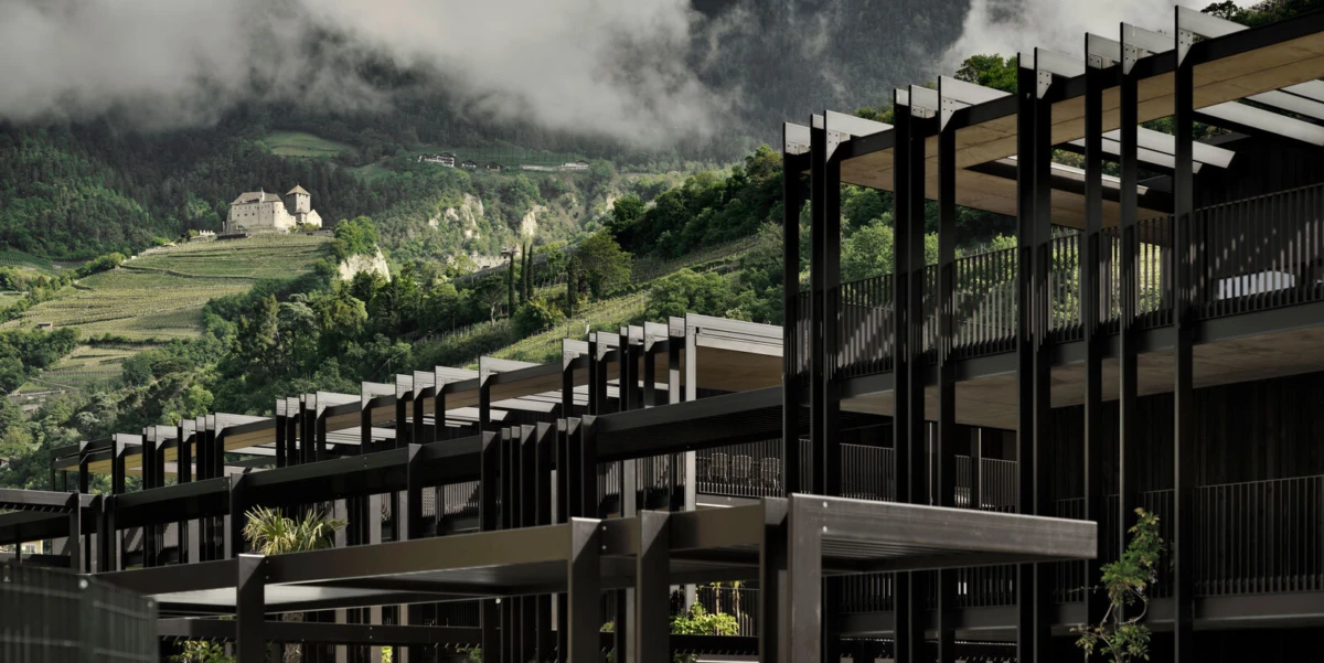 Modern black steel-framed building complex with open terraces stands beside hillside vineyards and a distant castle under cloudy sky.