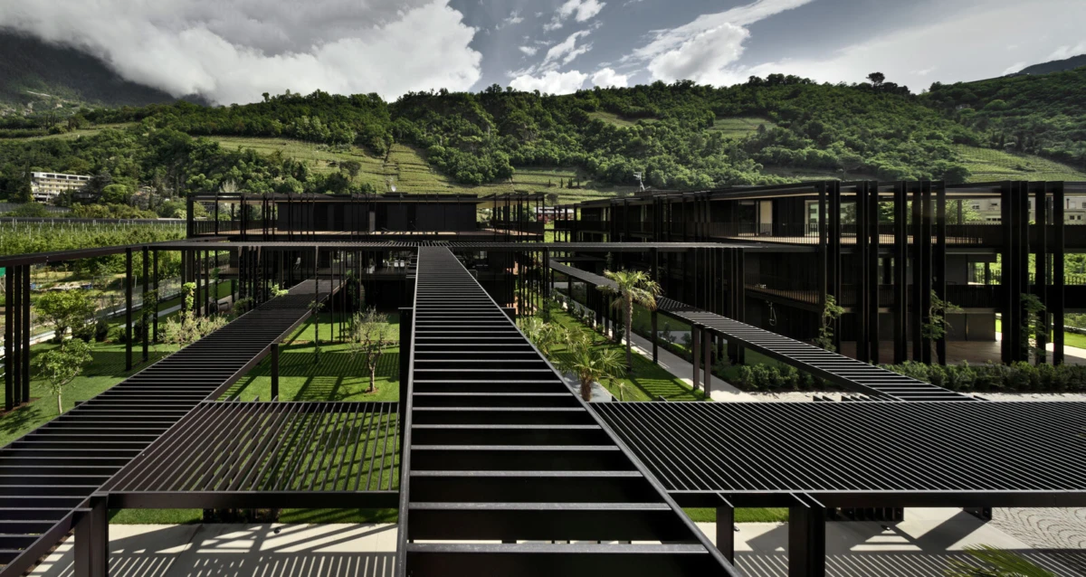 Aerial view of a steel-framed architectural complex with elevated slatted walkways and green courtyards, hillside landscape in background.