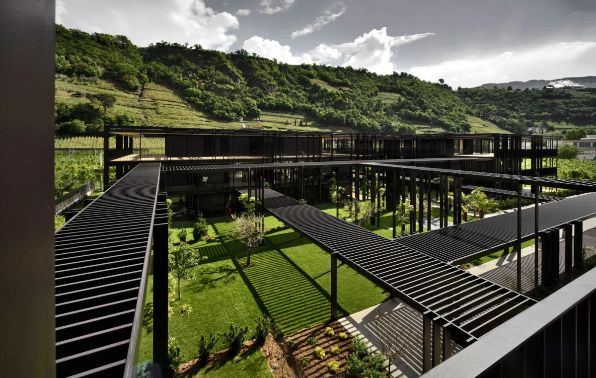 Modern steel-framed complex with elevated, crisscrossing walkways over a green courtyard; hillside backdrop.