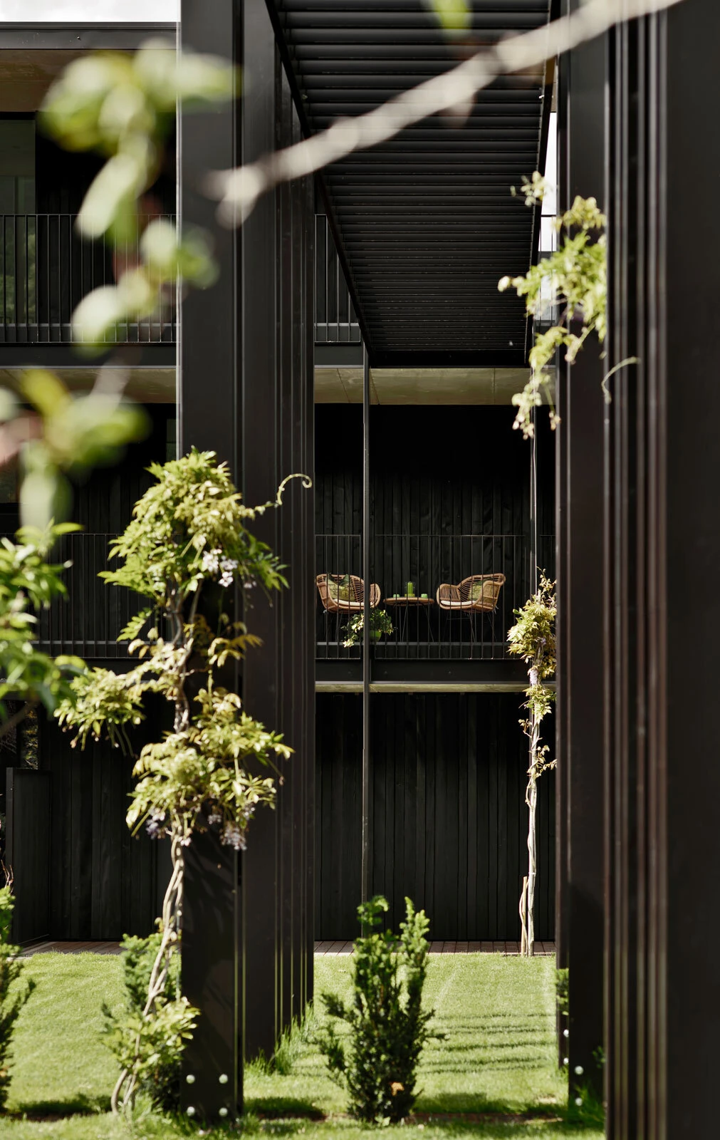 Two wicker chairs and a small table on a second-floor balcony of a dark modern building, overlooking a green courtyard.