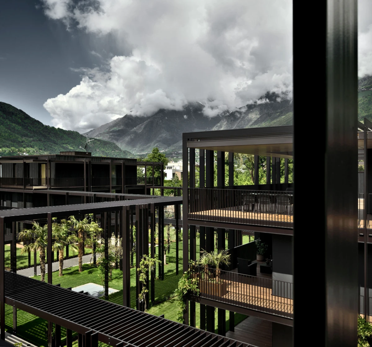 Modern black-metal residential complex with open walkways and balconies around a green courtyard; mountains and clouds in the distance.