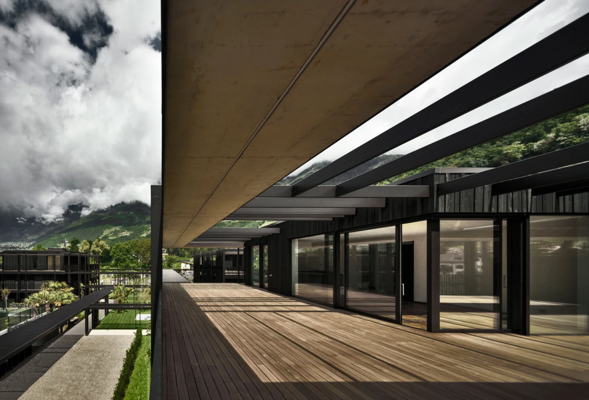 Contemporary glass-walled building with a long wooden deck terrace, empty; mountains and cloudy sky in the distance.