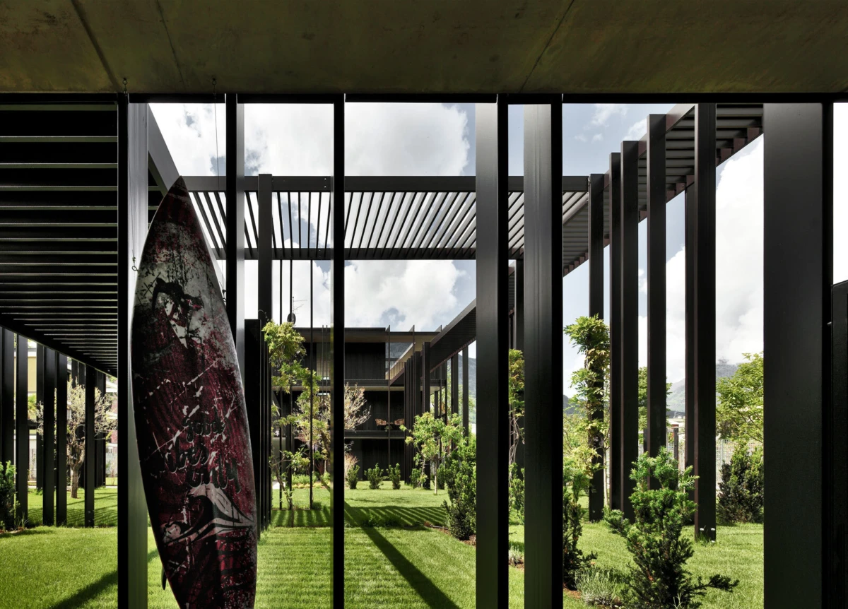 Red-patterned surfboard leaning against a black metal column in a modern courtyard with green lawn and trees.