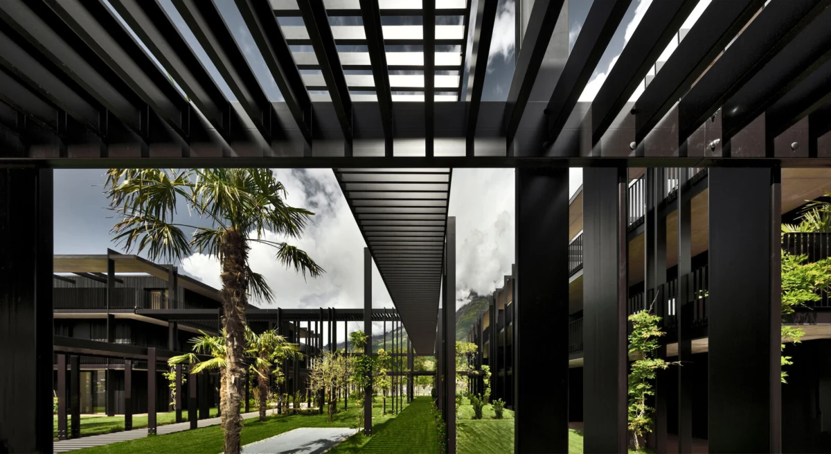Modern architectural courtyard with black steel columns and slatted overhead beams, palm trees, and a green lawn between dark buildings.