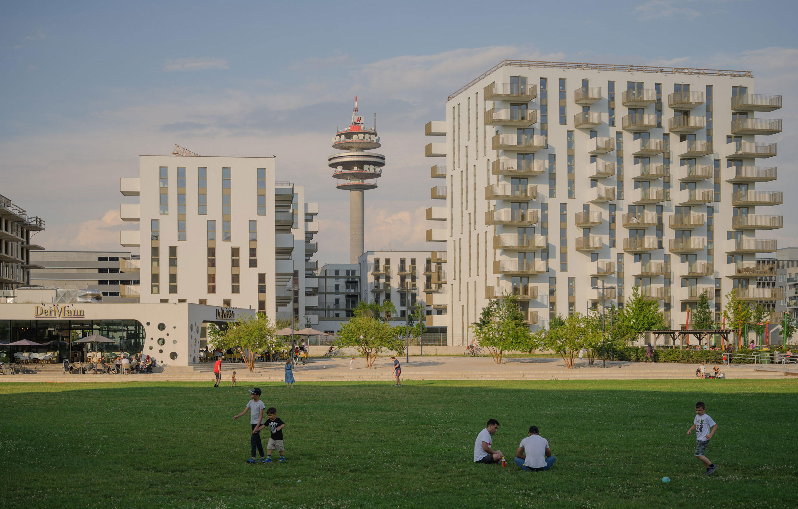Wide park in front of modern white apartment buildings; people walk, sit and play on the grass with a tall observation tower in the background.