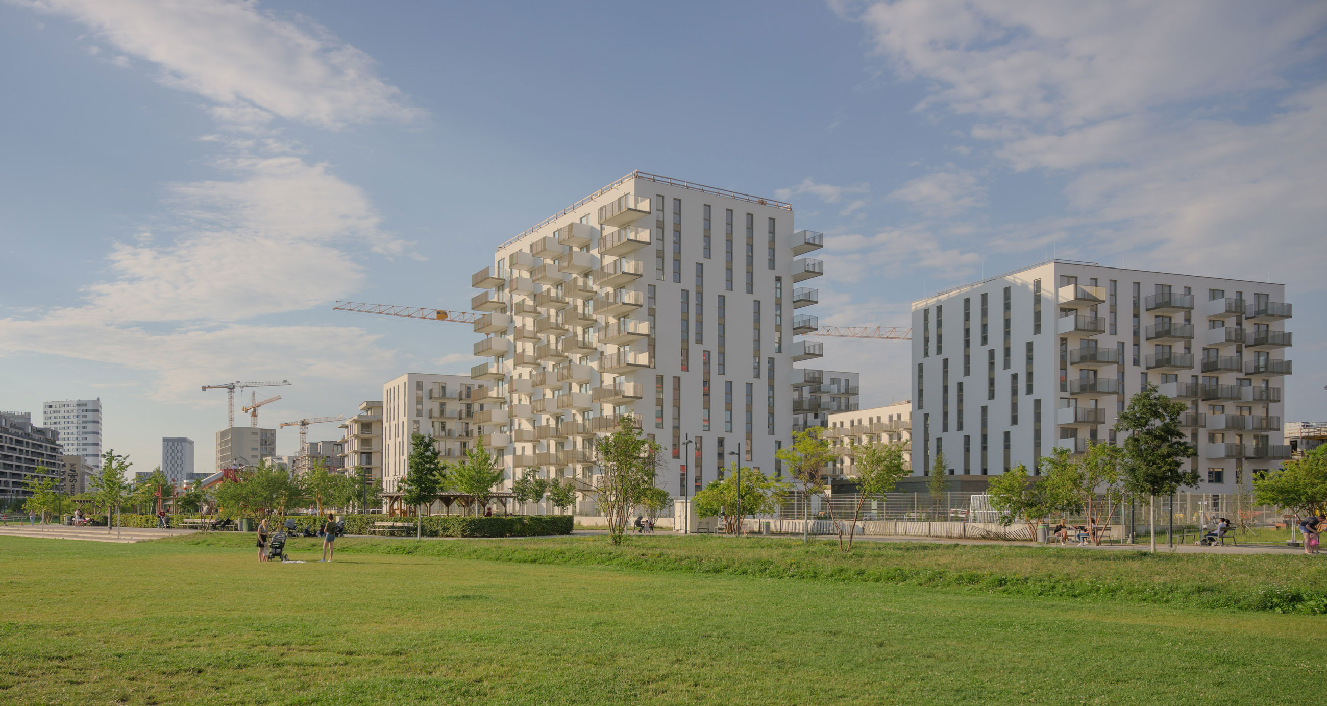 Modern white residential buildings under construction with cranes, set in a green park with pedestrians.