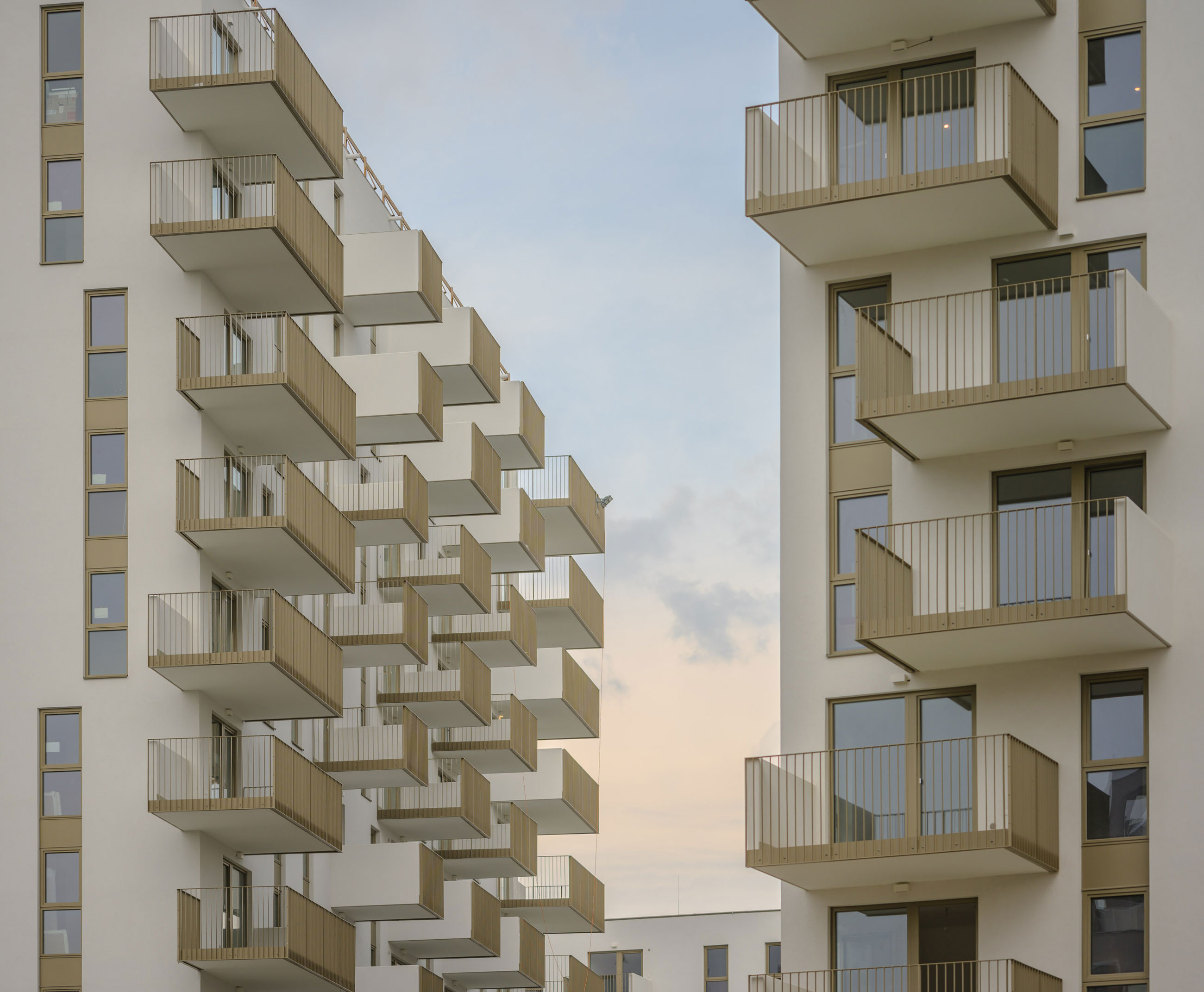 two white high-rise buildings with beige balconies arranged in stepped patterns, facing each other under a pale sunset sky.