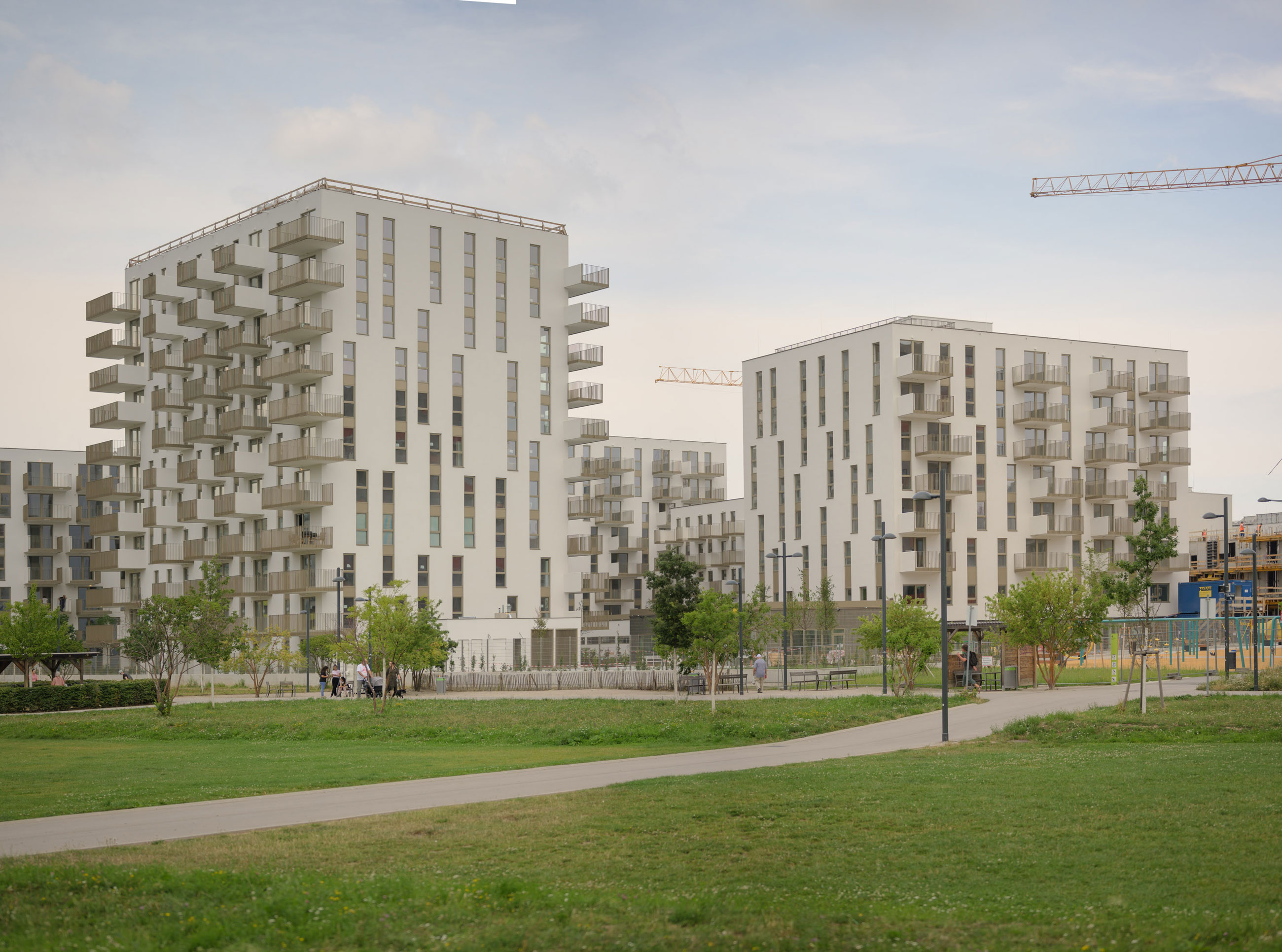 Modern apartment complex with staggered balconies under construction; cranes overhead, in a green park with a walking path.