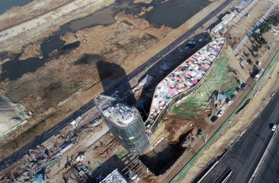 Construction site with a curved, multicolored-roofed building and a cylindrical tower beside a road and canal.