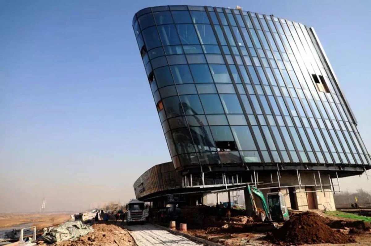 Leaning curved-glass skyscraper under construction on a dirt site with heavy machinery nearby.