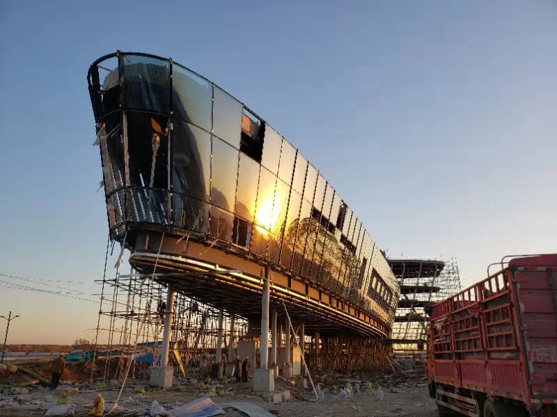 Curved glass structure on elevated stilts under construction; scaffolding surrounds a debris-filled site, with a red truck nearby.