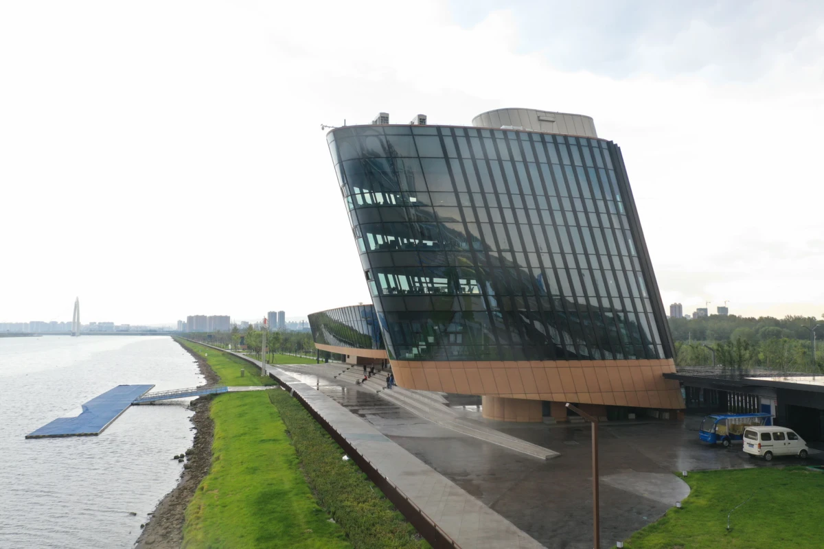 Glass-clad, diagonally tilted building by the river, with a riverside promenade, green lawn, and parked vans nearby.