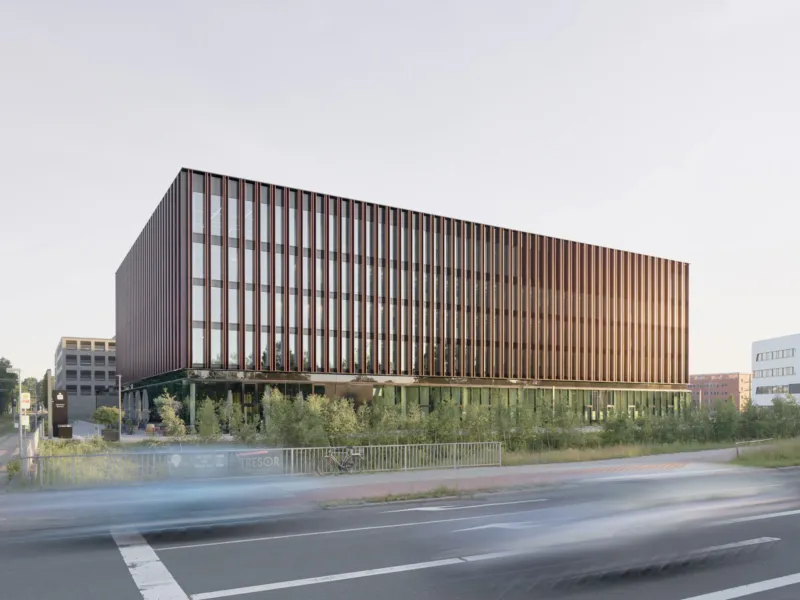 Glass-fronted office building with vertical brown fins beside a street; motion-blurred cars pass as greenery lines the foreground.