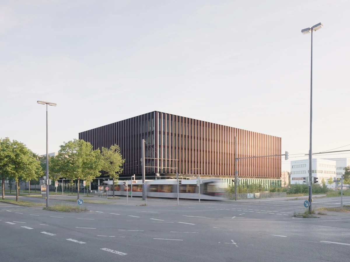 Glass-clad office building with vertical fins at a busy intersection; a motion-blurred tram runs past, with trees and streetlights along the roadside.