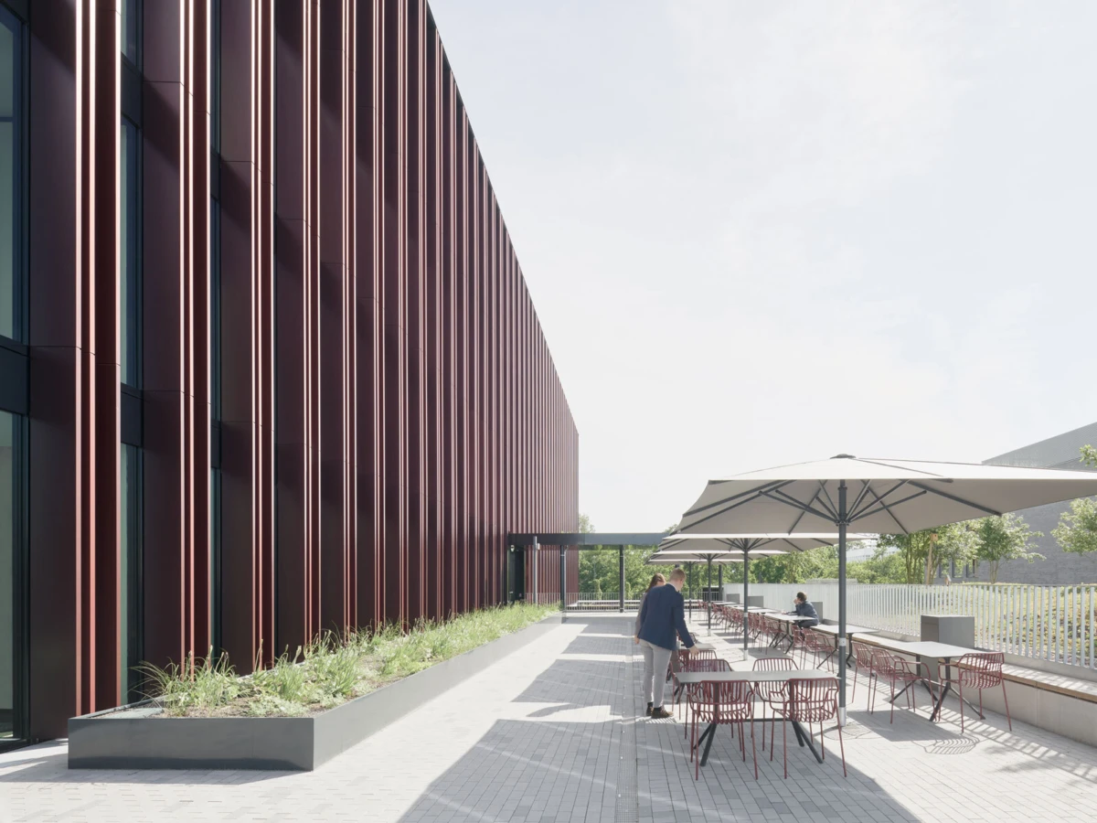 Outdoor cafe terrace beside a modern red-metal facade with vertical fins; a person stands by a table under umbrellas.