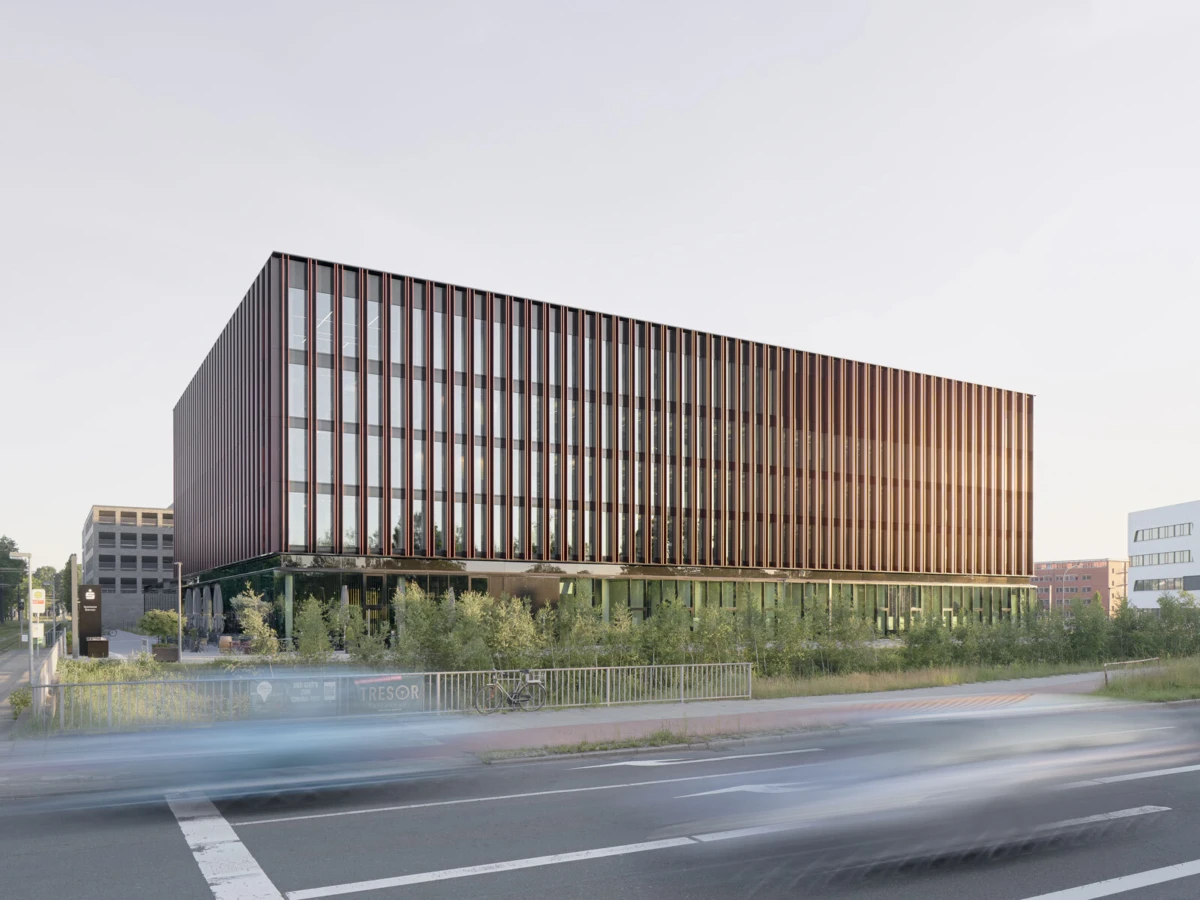 Glass-fronted office building with vertical brown fins beside a street; motion-blurred cars pass as greenery lines the foreground.