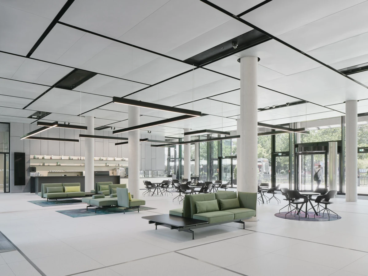 Open-plan office lobby with white columns, green modular sofas arranged for lounging, black chairs, and large glass doors.