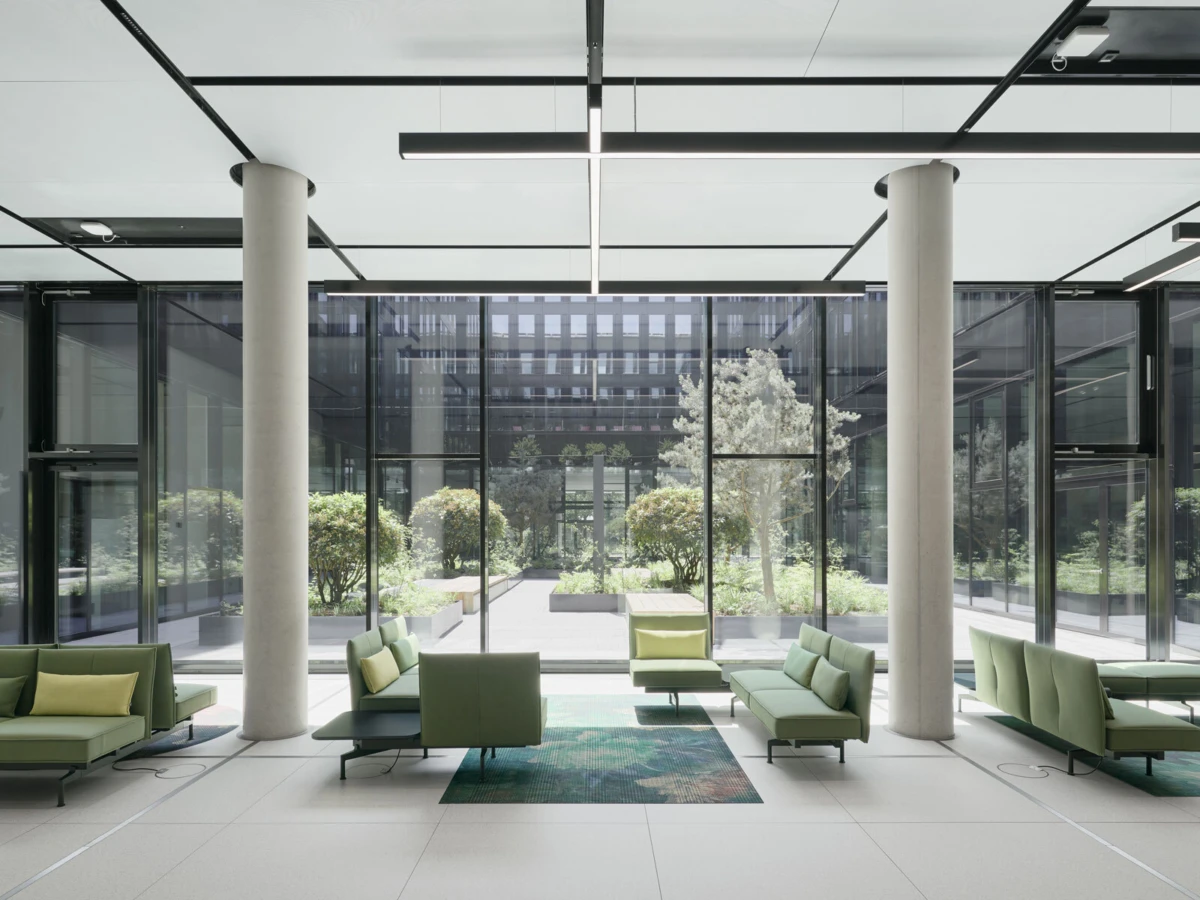Empty modern glass-walled atrium lounge with green sectional sofas, low tables, and a central rug; trees visible through windows.
