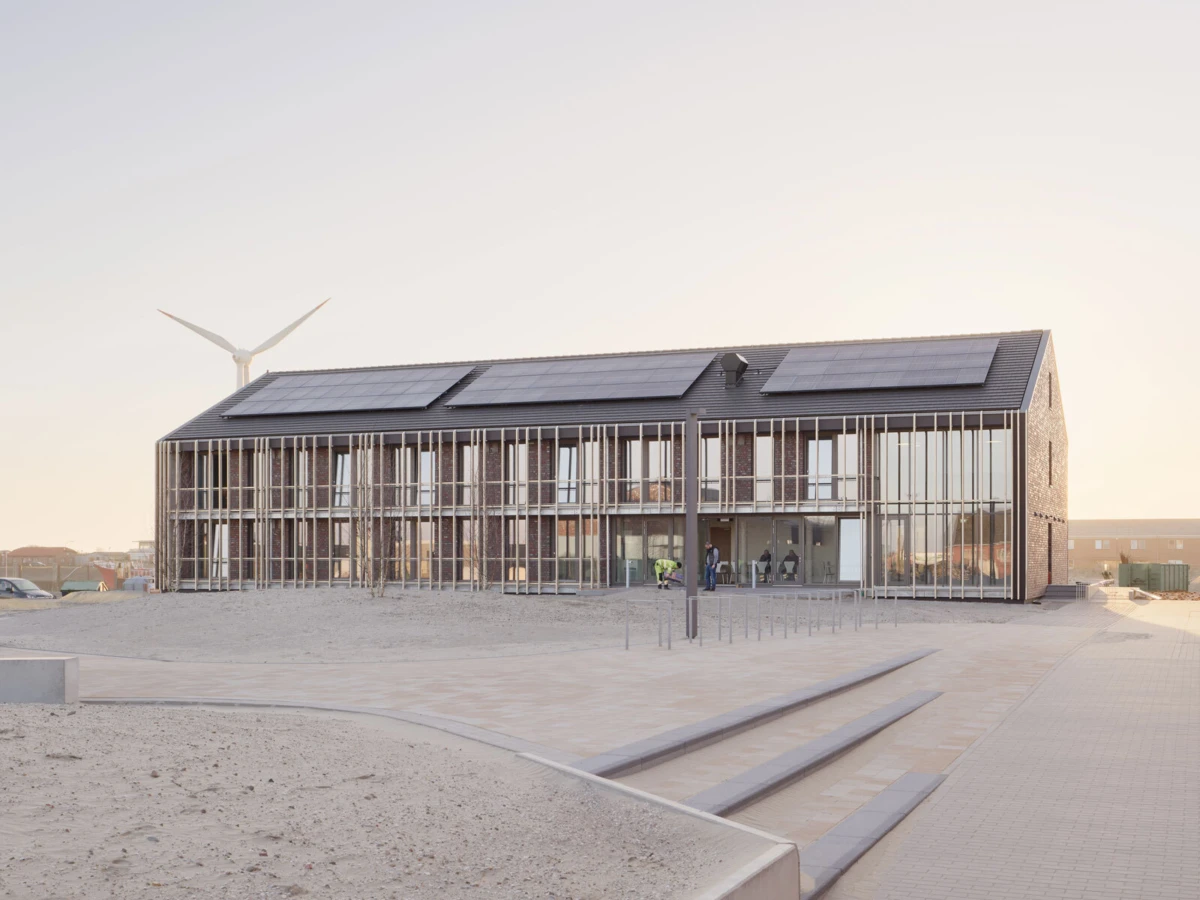 Modern beachside pavilion with glass walls, vertical slats, and roof solar panels; wind turbine in the distance.