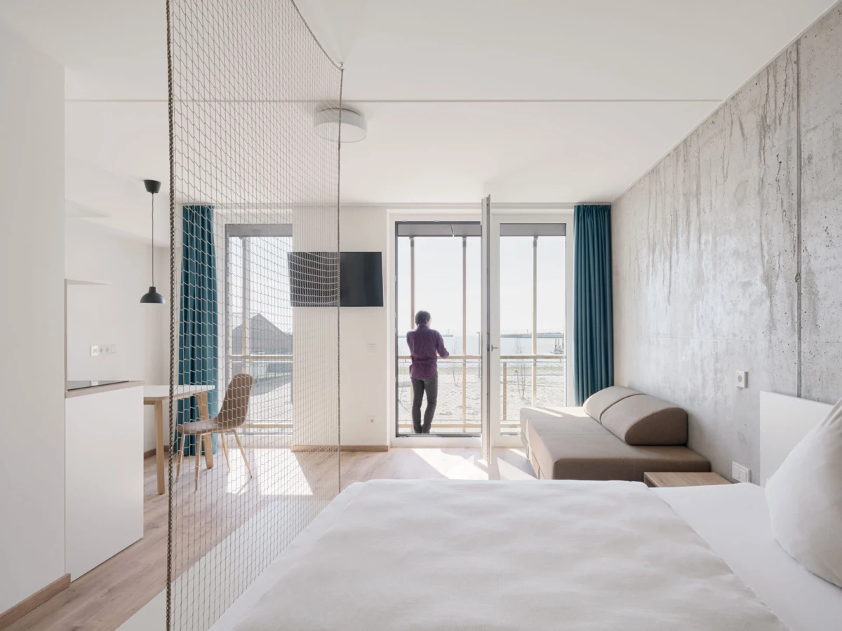 Person stands on the balcony of a modern hotel room, with a bed, sofa, and mesh divider, overlooking a seascape.