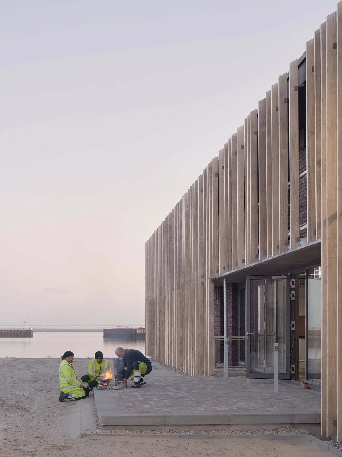 Four construction workers in hi-vis jackets circle a small campfire on a paved waterfront plaza beside a wooden-clad building.