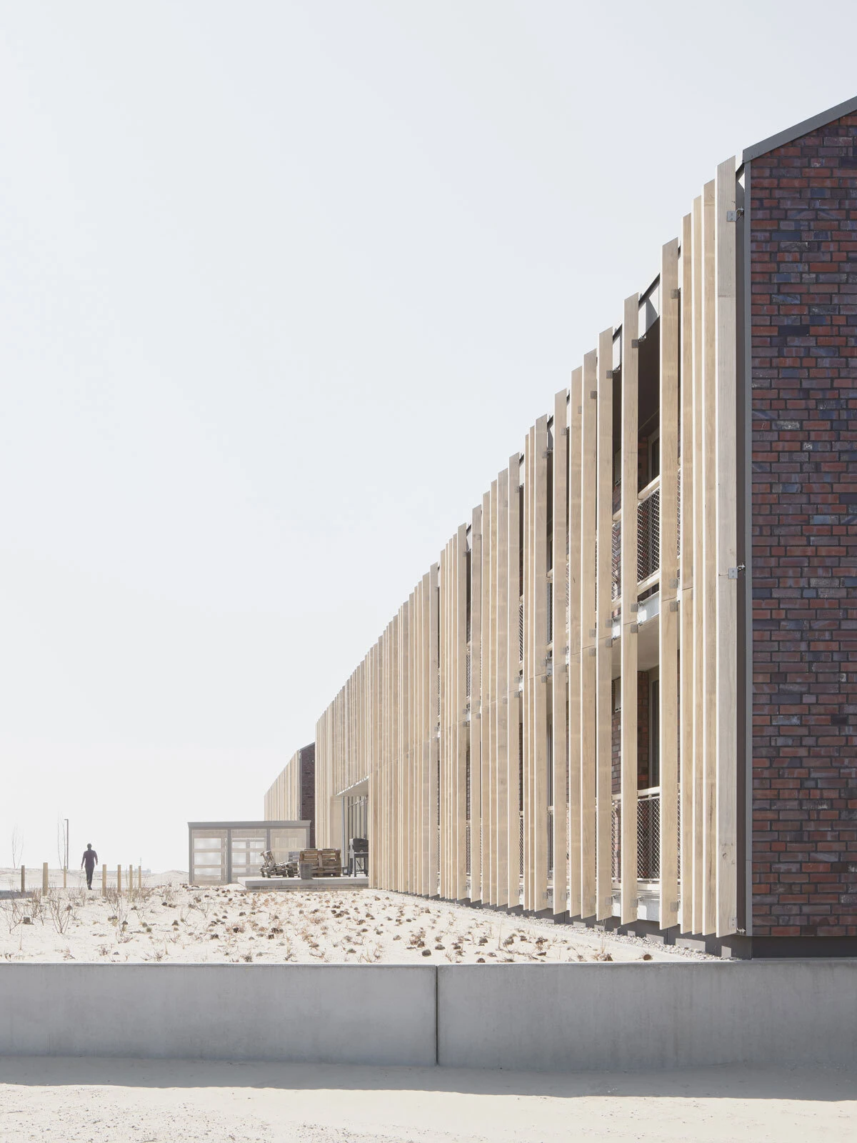 Modern brick-and-wood building with tall vertical wooden slats along a sandy coastal lot; a distant figure walks.