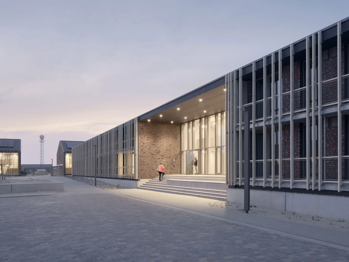 Exterior view of a modern brick-and-glass building with metal louvers; a person walks up the steps toward the glass entrance at dusk