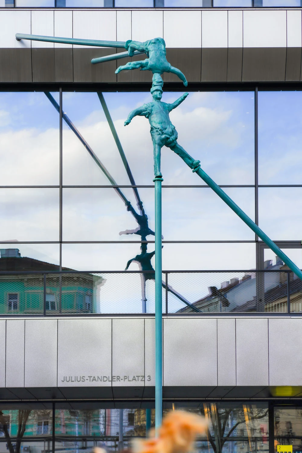 Turquoise sculpture of two acrobats balancing on a vertical pole in front of a glass-fronted building.