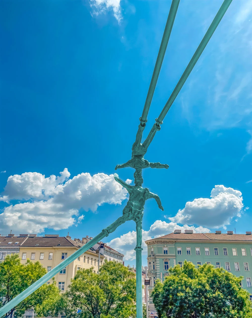Metal sculpture of human figures clinging to a vertical pole, suspended by diagonal cables in a sunny urban square.