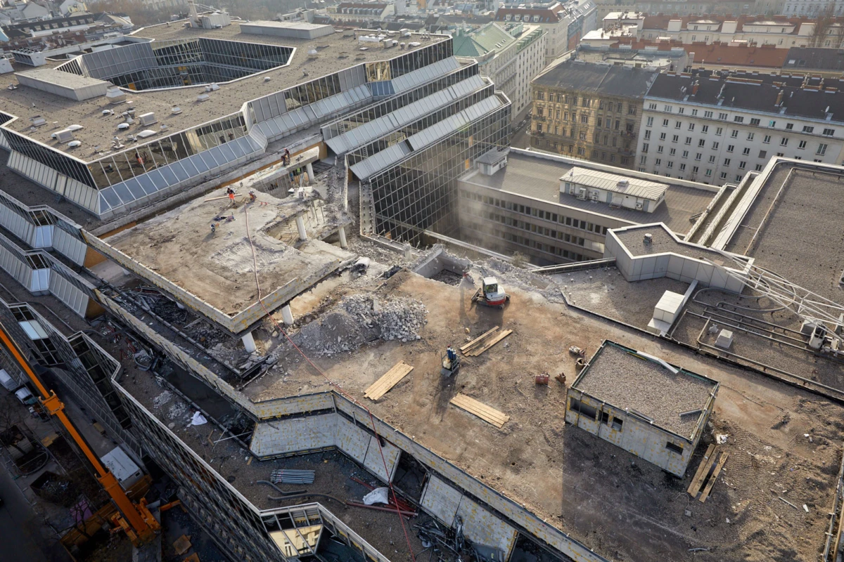 Aerial view of a rooftop construction site with demolition rubble, workers, and machinery amid a dense urban cityscape.