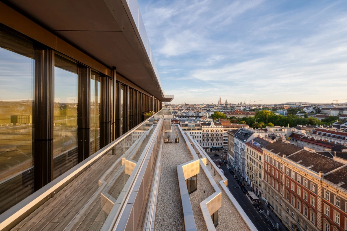 Glass-walled rooftop walkway along a modern building, overlooking a city at sunset.