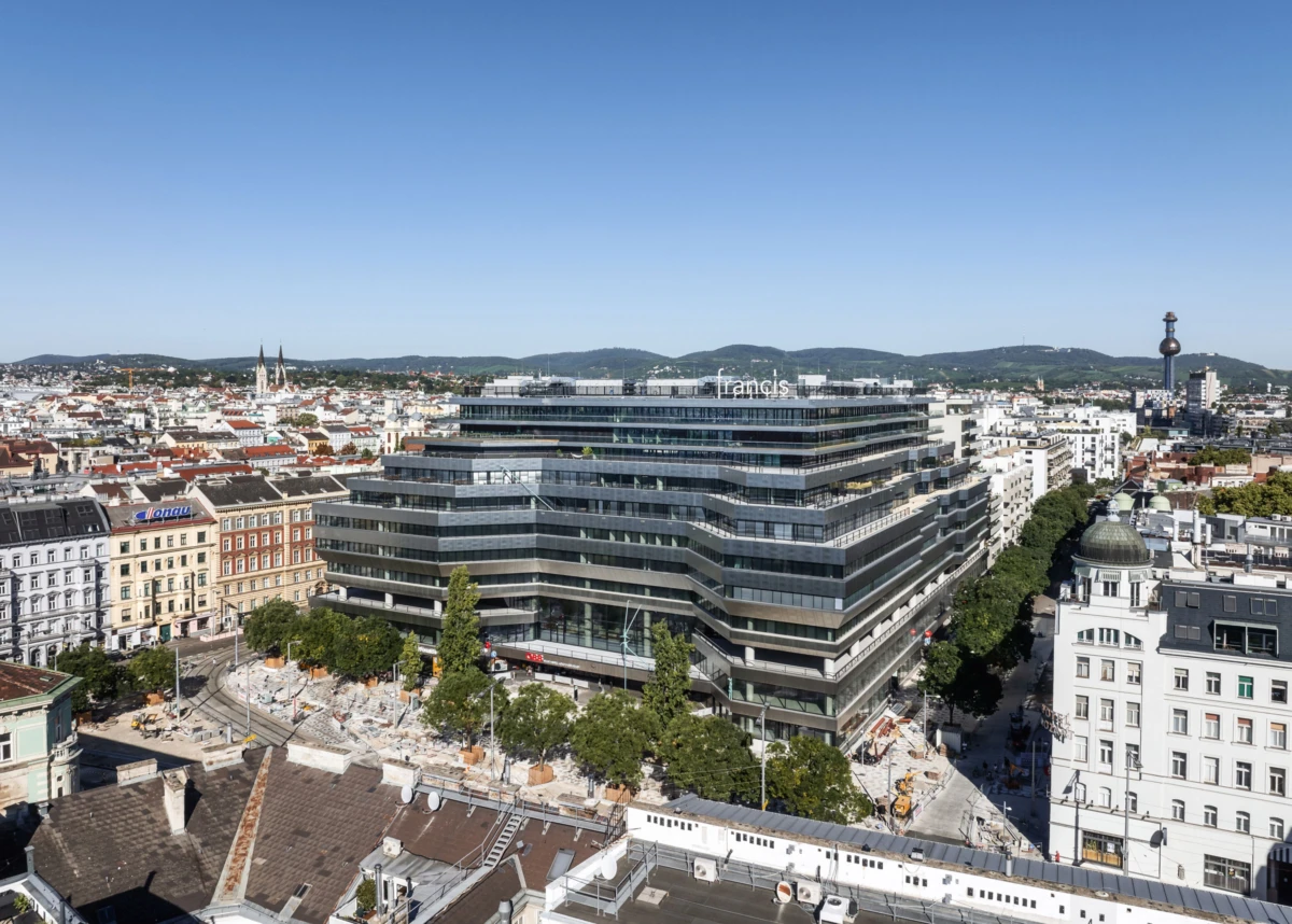 Dominant tiered glass office building with francis sign, seen in an aerial daytime view over historic European streets.