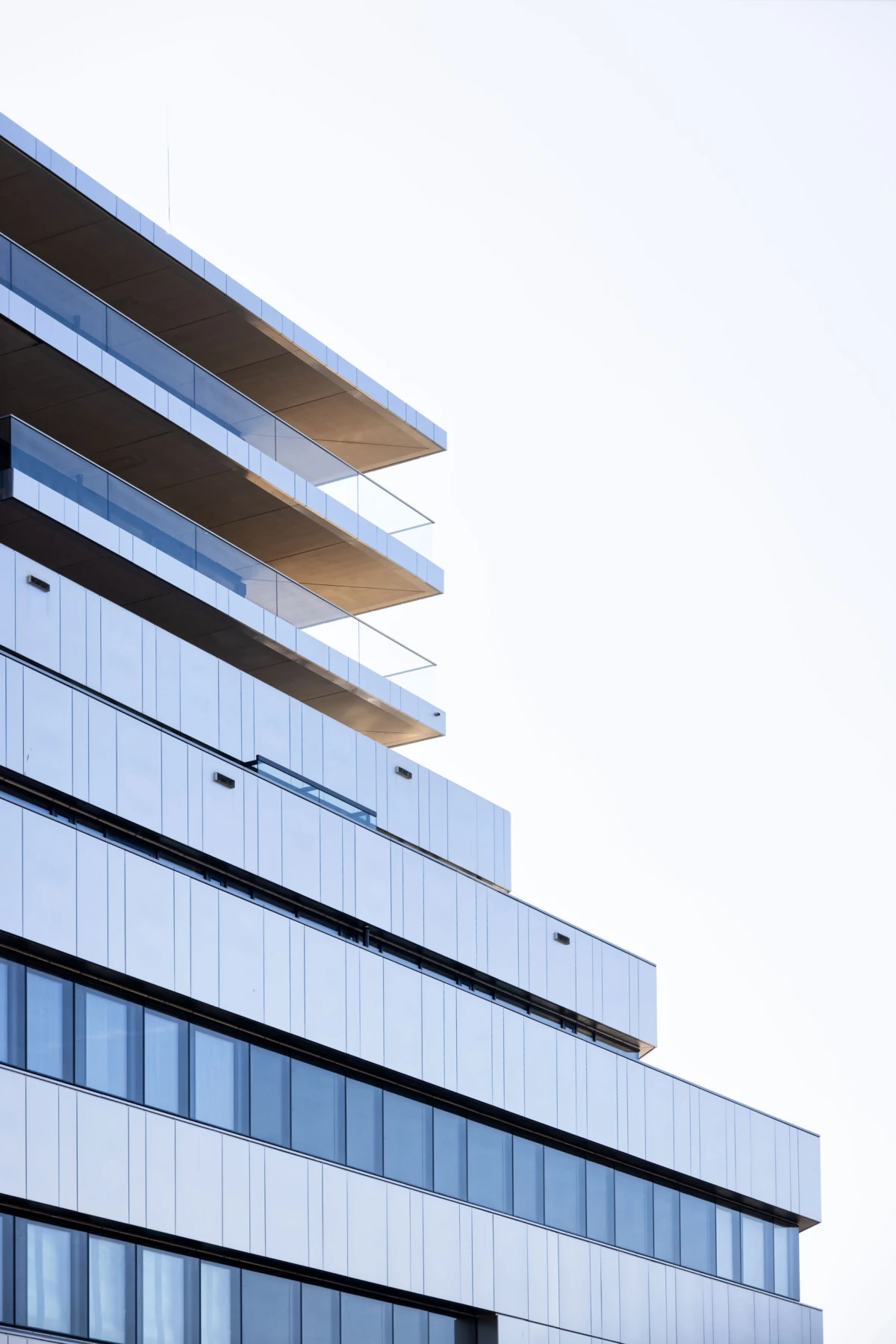 Modern glass-and-steel building with stacked, protruding balconies on the left and blue-tinted windows against a bright sky.