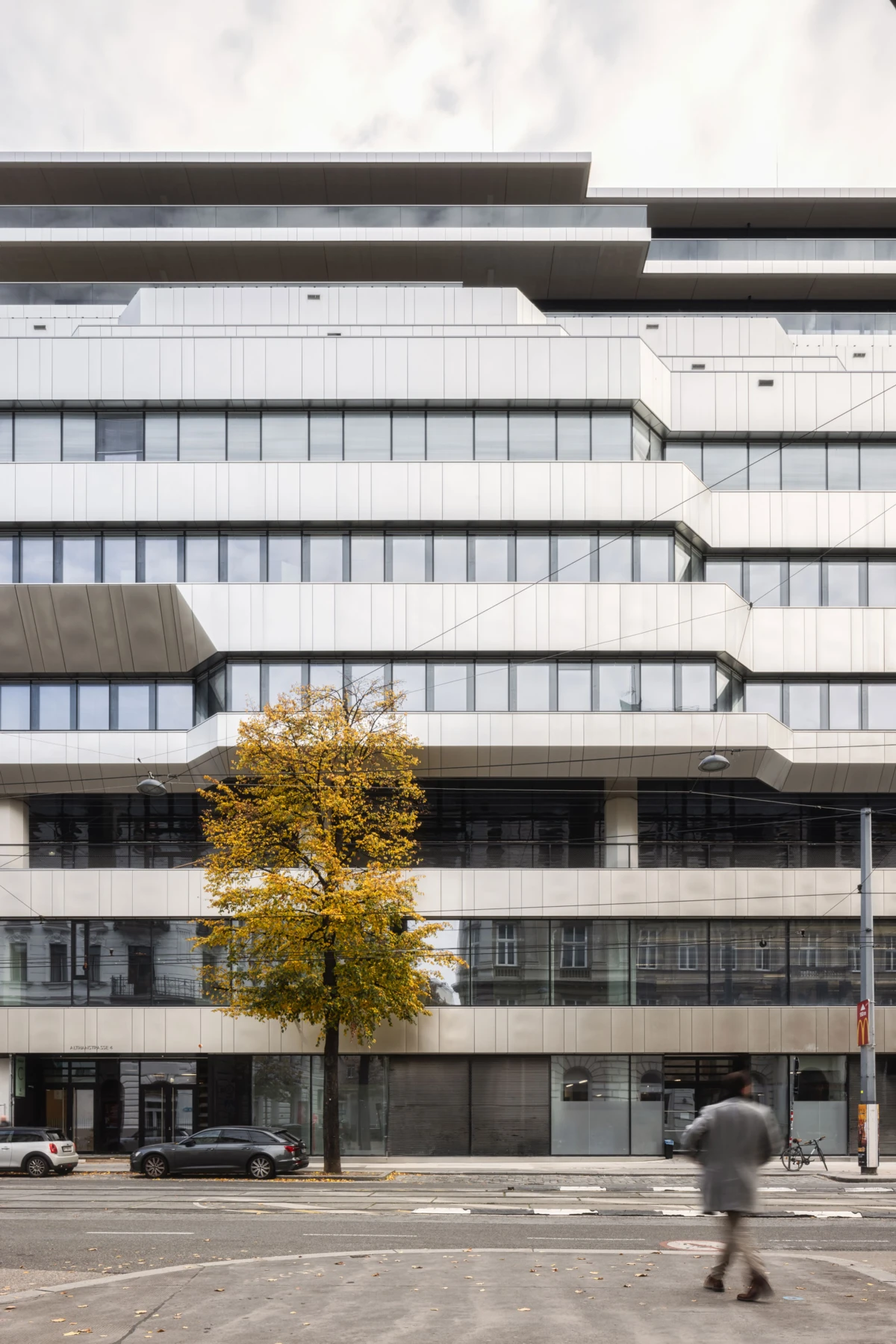 Modern tiered office building with staggered facades; yellow autumn tree in front; pedestrian walking along a city street.
