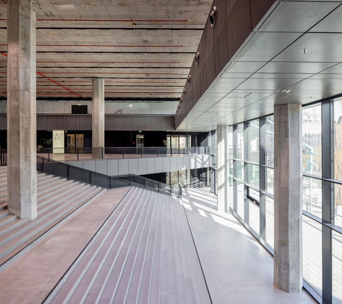Empty modern atrium with tiered concrete seating, tall pillars, and floor-to-ceiling glass walls.