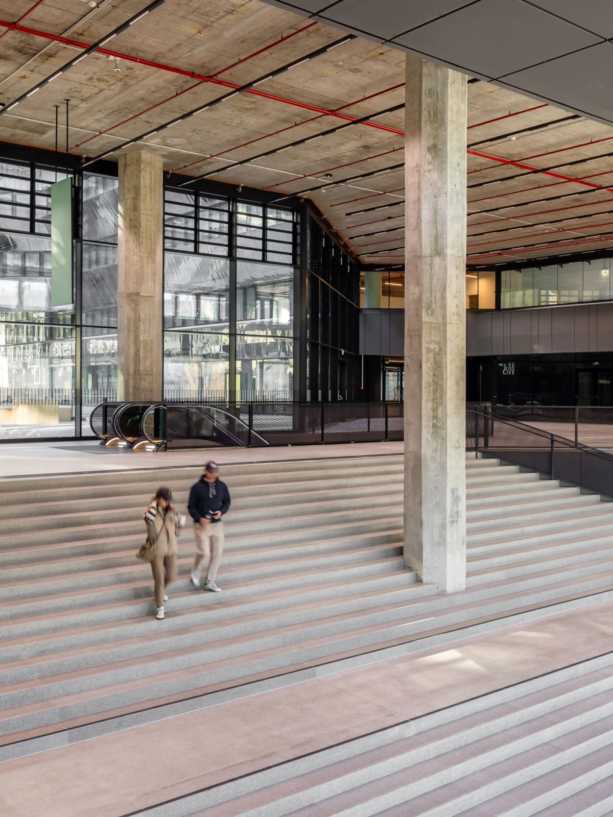 Two pedestrians descend wide steps inside a modern atrium with concrete pillars and glass walls.