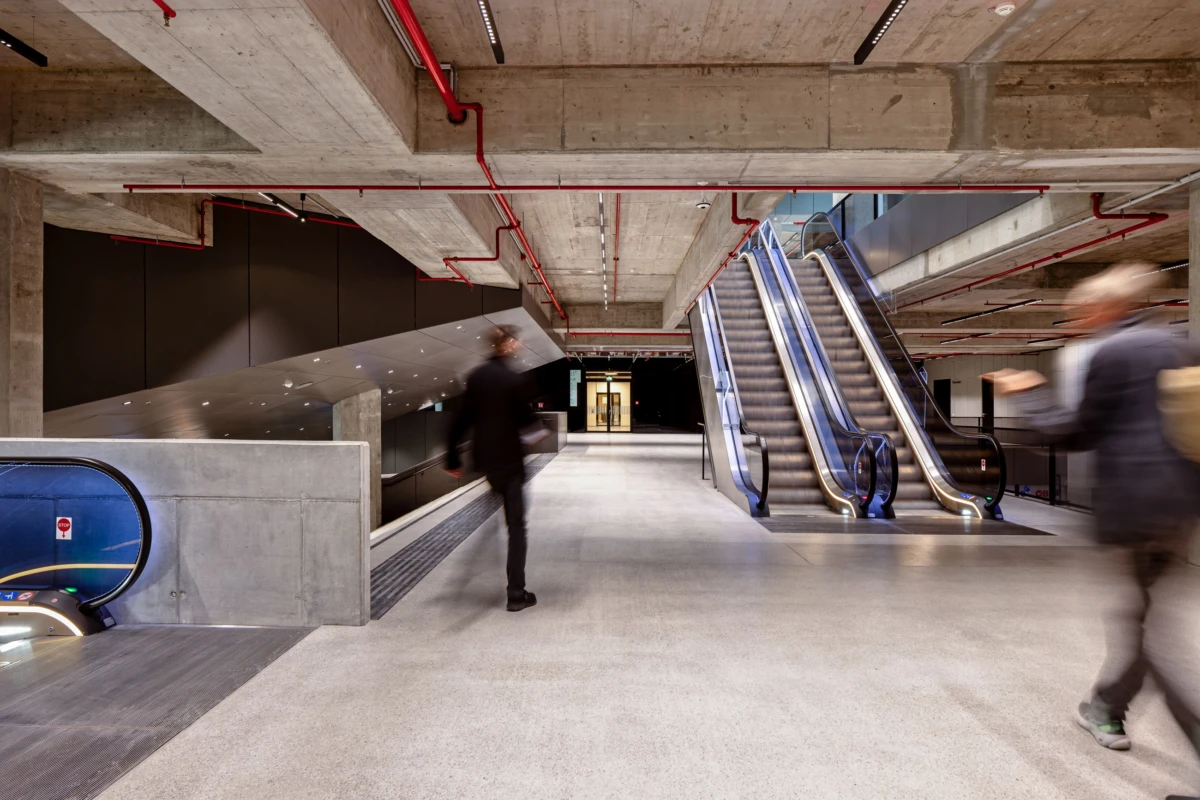 Two escalators in a concrete, industrial transit concourse; blurred pedestrians walking by.