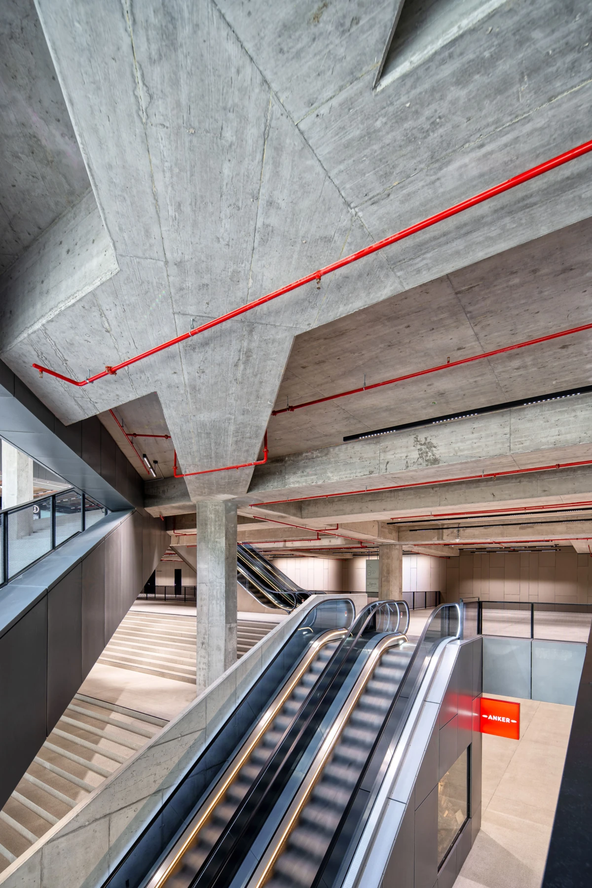 Two parallel escalators in a modern concrete atrium with exposed red pipes along the ceiling.