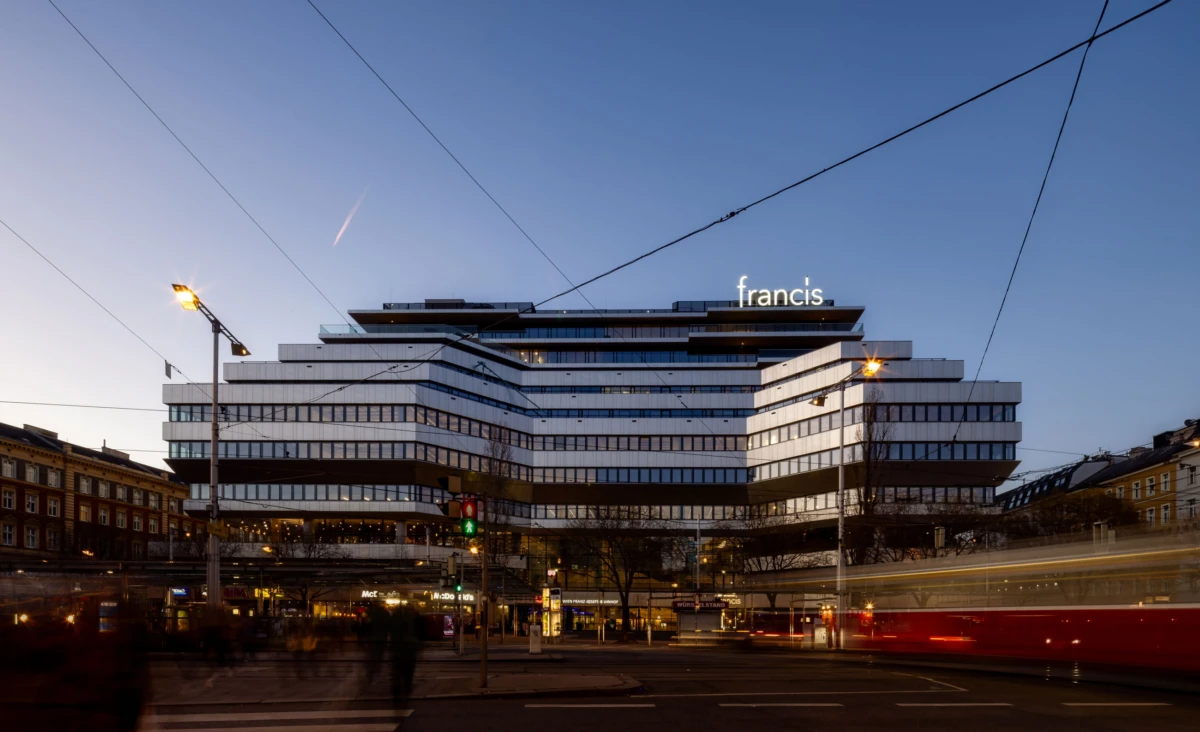 Modern tiered office building with a prominent 'francis' sign at dusk, as traffic streaks cross a city intersection in front.