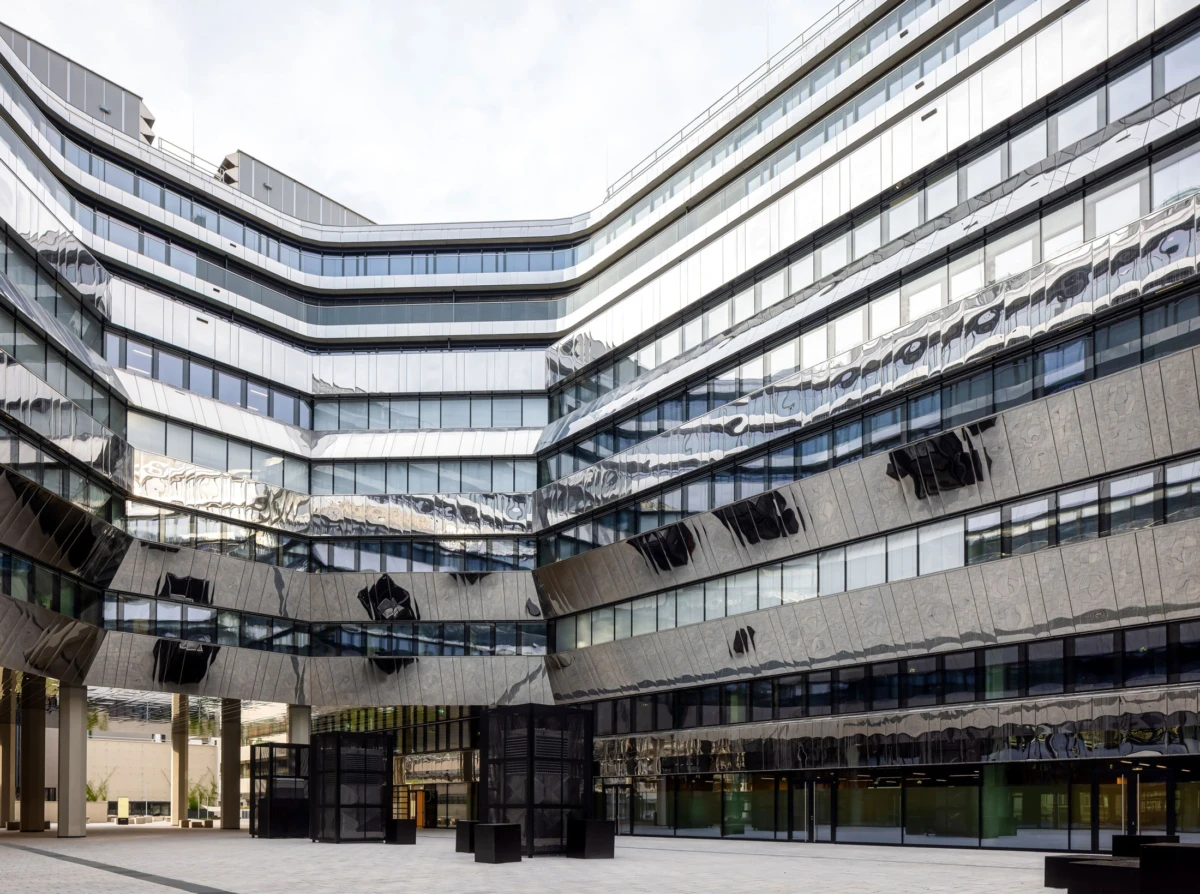 Modern glass office building with angular, stacked façades and mirrored surfaces surrounding a paved courtyard with black planters.