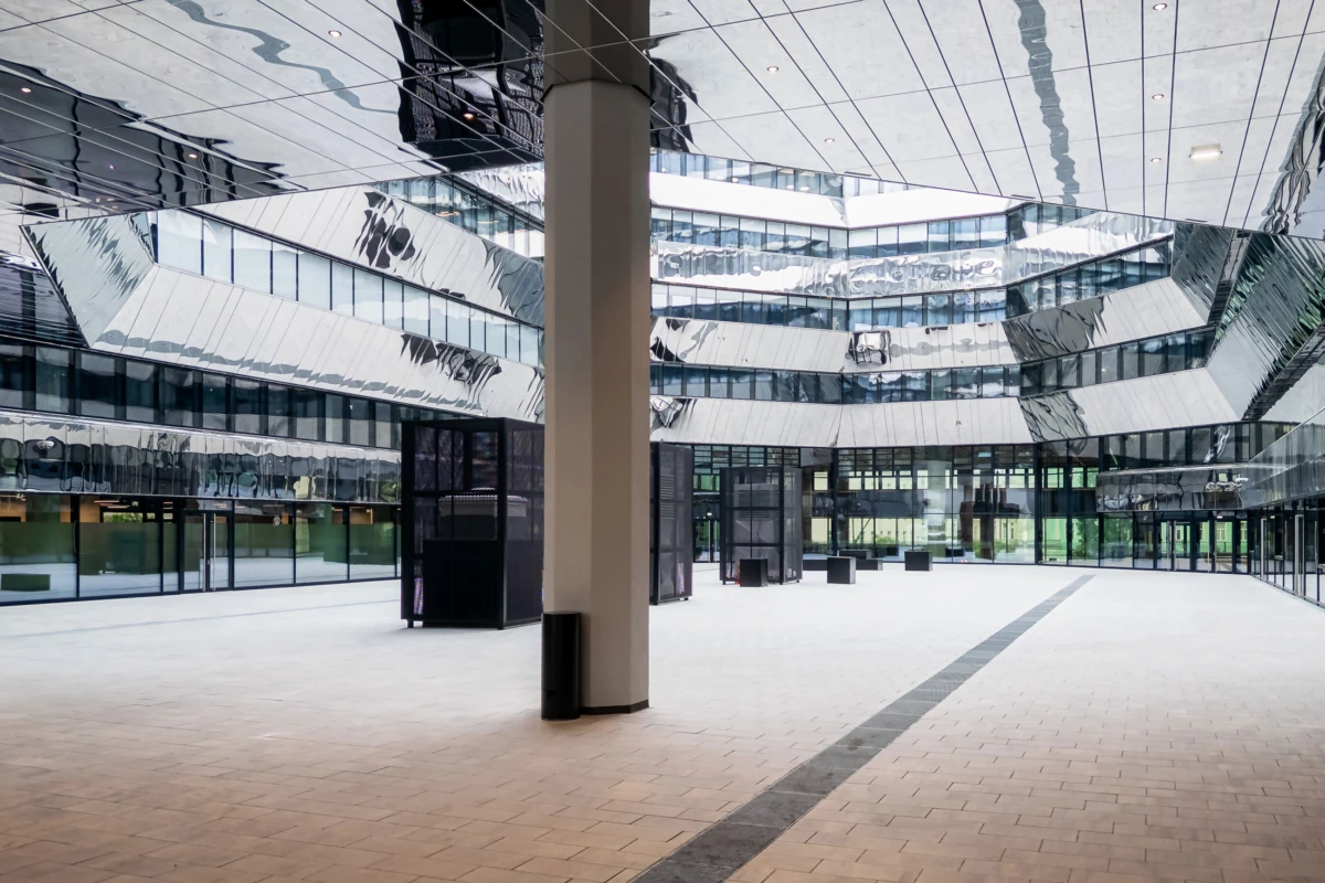 Empty modern glass atrium courtyard with a reflective metal facade and angular surrounding buildings over a wide tiled plaza.