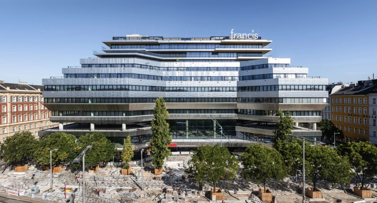 Multi-tier glass office building with a Francis sign atop, towering over a city square with trees and tram tracks.
