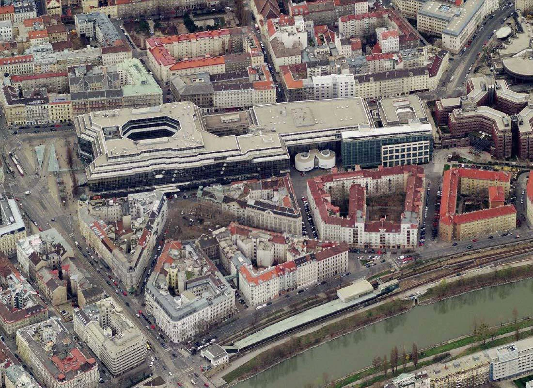 Aerial view of a dense city center with a large modern office complex near a winding river, surrounded by red-roofed buildings.