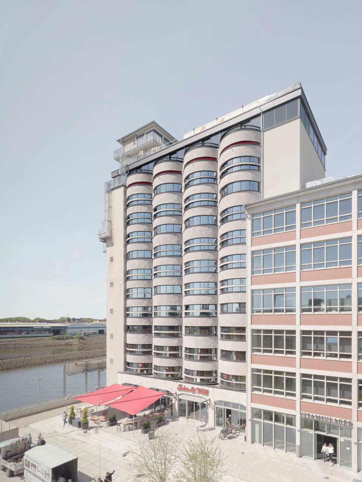 Tall cylindrical residential tower with curved balconies beside a river, ground-level cafe under a red canopy.